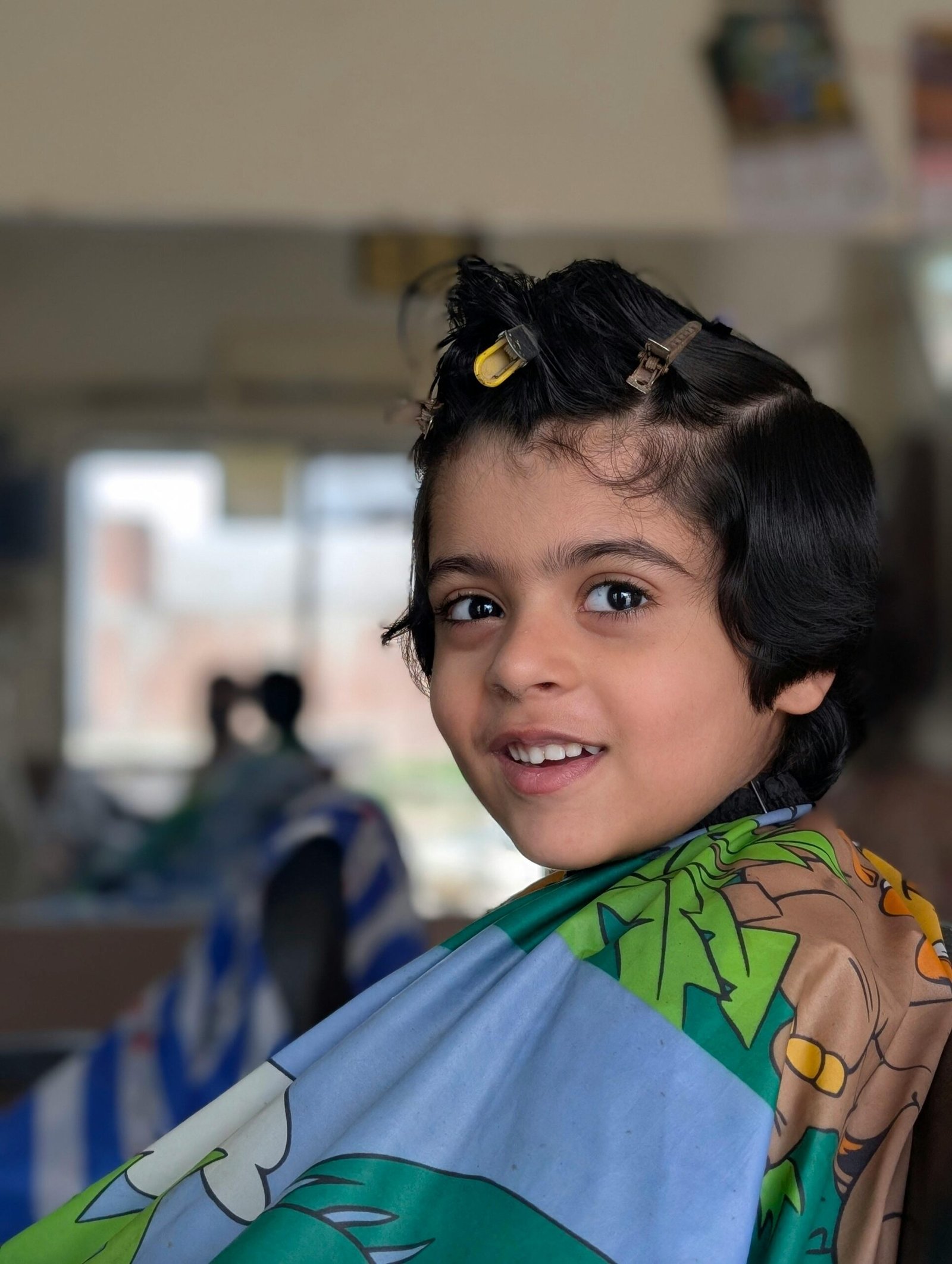 A young child at a salon in Pakistan smiling while getting a haircut wearing a colorful cape.